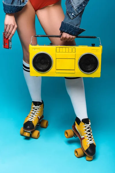 Cropped view of woman in swimsuit and roller skates holding boombox and cassette tape on blue — Stock Photo