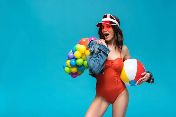 Chica excitada en traje de baño y patines de ruedas celebración de pelota de playa y bolsa de picadura con bolas de colores aislados en azul - foto de stock