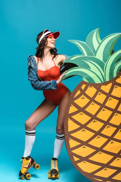 Vue pleine longueur de la femme en maillot de bain et patins à roulettes debout près de l'ananas décoratif sur bleu — Photo de stock