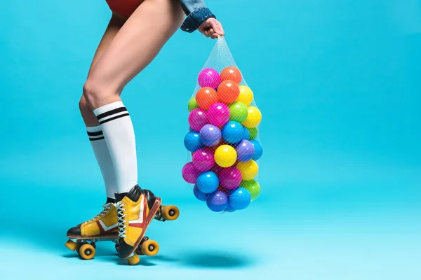 Cropped view of woman in knee socks and roller skates holding string bag with colorful balls on blue — Stock Photo