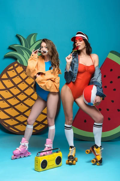 Full length view of two stylish girls in roller skates with beach ball and armelon lollipop on blue — стоковое фото