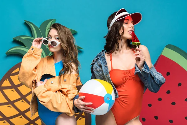 Duas meninas com bola de praia e pirulito perto de melancia decorativa e abacaxi isolado em azul — Fotografia de Stock