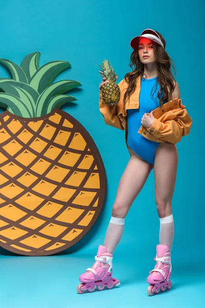 Full length view of girl in swimsuit and roller skates holding pineapple on blue — Stock Photo