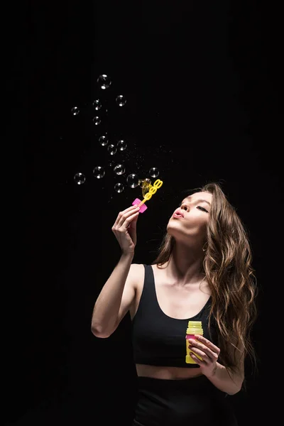 Красивая молодая женщина выдувает мыльные пузыри изолированные на черном — стоковое фото