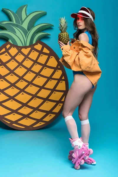 Vue pleine longueur de fille sexy en patins à roulettes tenant l'ananas sur bleu — Photo de stock