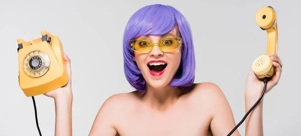 Возбужденная обнажённая женщина в фиолетовом парике держит ретро-телефон, изолированный на сером — стоковое фото