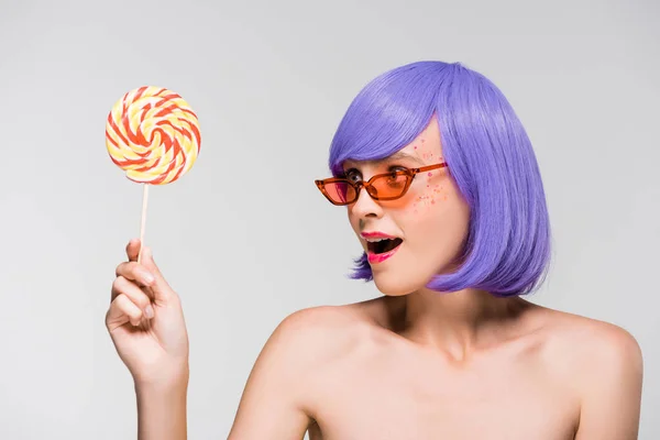 Surprised girl in purple wig and sunglasses looking at lollipop, isolated on grey — Stock Photo