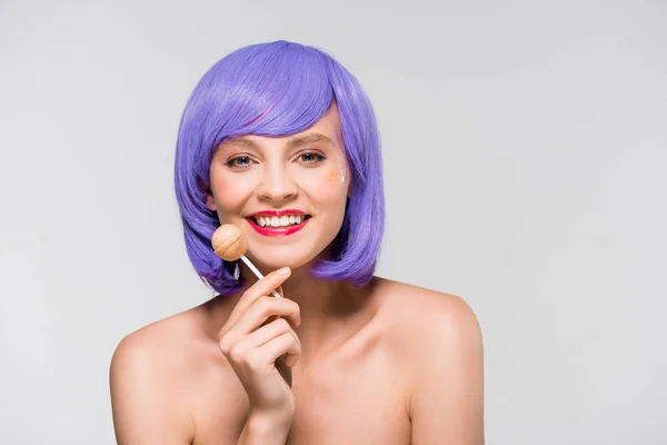 Chica sonriente en peluca púrpura sosteniendo dulce piruleta, aislado en gris — Stock Photo