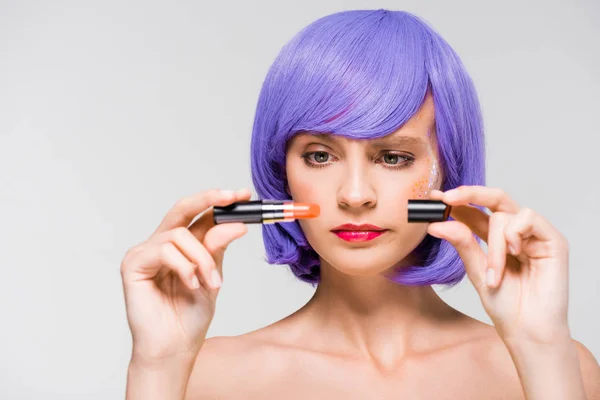 Confused girl in purple wig holding lipstick isolated on grey — Stock Photo