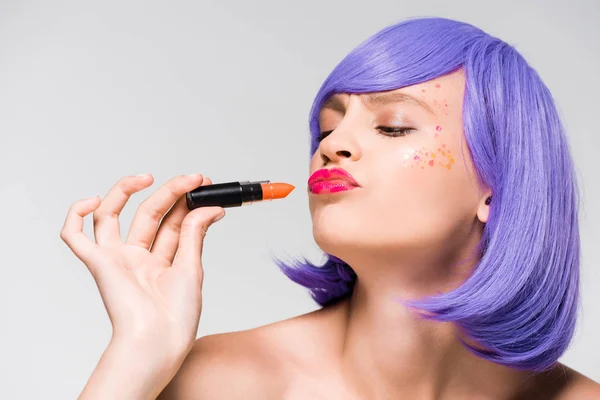 Elegante hermosa chica en peluca púrpura celebración de lápiz labial aislado en gris - foto de stock