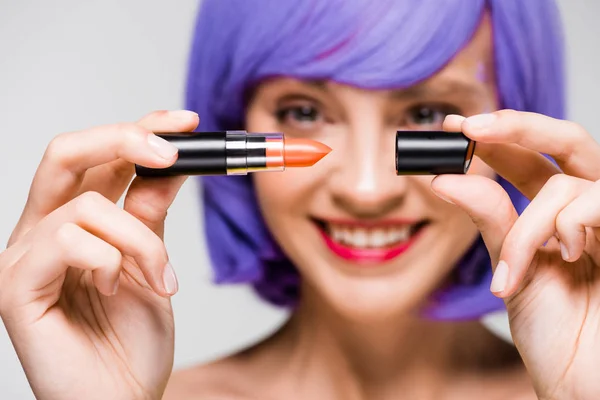 Selective focus of smiling girl in purple wig holding lipstick isolated on grey — Stock Photo