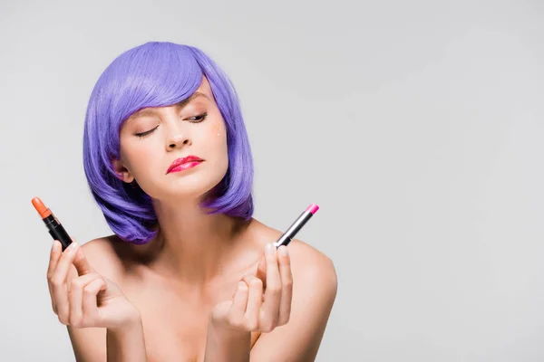 Jeune femme réfléchie en perruque violette regardant les rouges à lèvres isolés sur gris — Photo de stock