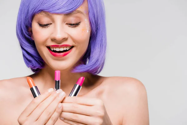 Hermosa mujer sonriente en peluca púrpura mirando lápices labiales aislados en gris - foto de stock