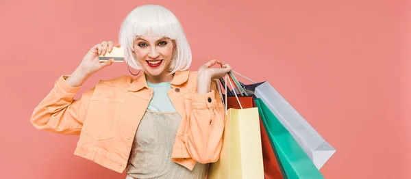 Улыбающаяся девушка в белом парике с пакетами покупок и кредиткой, изолированная на розовый — стоковое фото