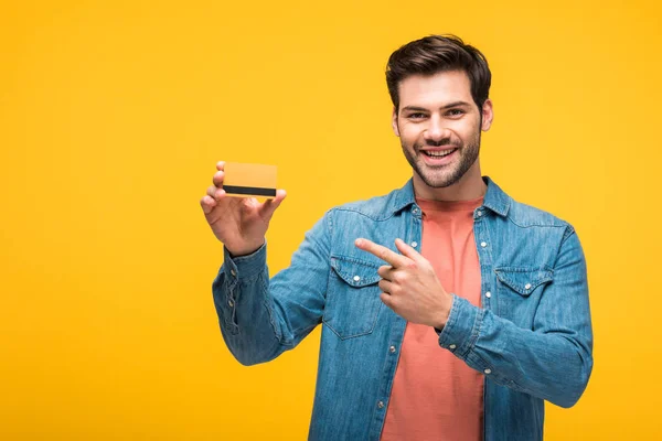 Feliz hombre guapo apuntando con el dedo a la tarjeta de crédito aislado en amarillo - foto de stock