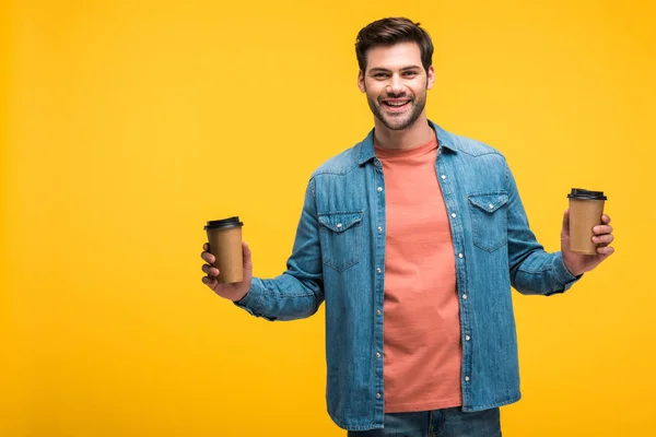 Bel homme souriant tenant des tasses en papier avec du café pour aller isolé sur jaune — Photo de stock