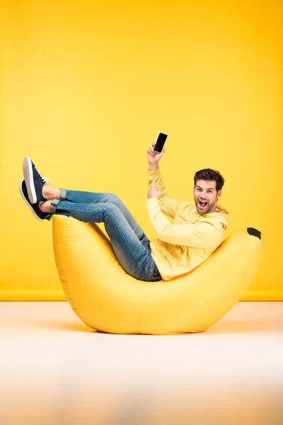 Homme heureux sur chaise sac haricot tenant smartphone sur jaune — Photo de stock
