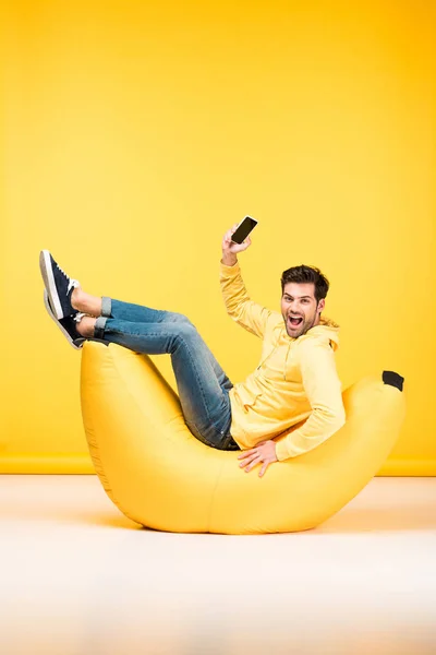 Homem feliz na cadeira de saco de feijão segurando smartphone no amarelo — Fotografia de Stock