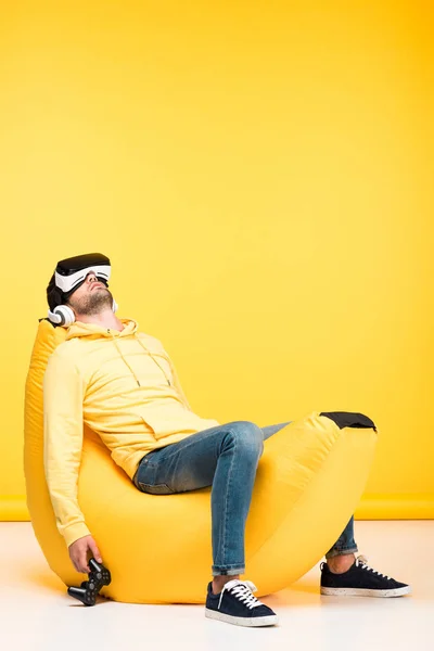 KYIV, UCRANIA - 12 DE ABRIL: hombre en la silla de la bolsa de frijol con joystick en auriculares de realidad virtual en amarillo - foto de stock