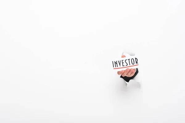 Visão parcial do homem segurando cartão com palavra investidor através de furo na parede de papel no branco — Fotografia de Stock