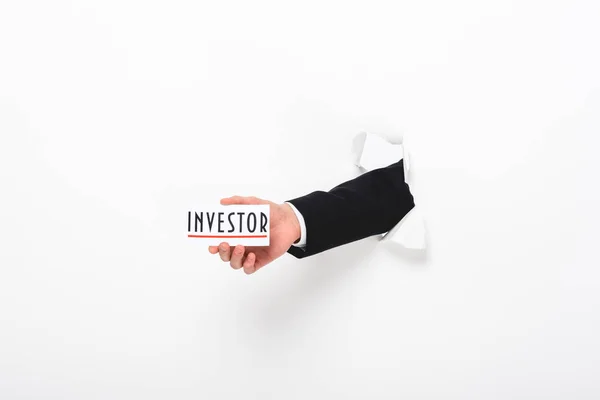 Cropped view of man holding card with investor word through hole in paper wall on white — Stock Photo