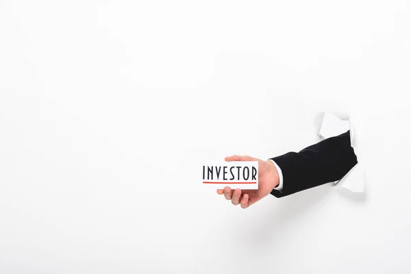 Cropped view of man holding card with investor word through hole in paper wall on white — Stock Photo