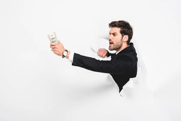 Angry businessman holding dollar banknotes behind hole in paper wall on white — Stock Photo