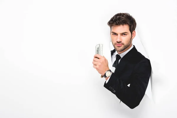 Handsome businessman holding dollar banknotes behind hole in paper wall on white — Stock Photo