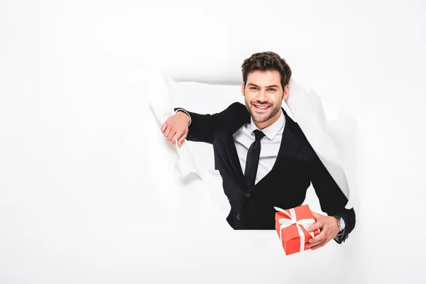 Uomo bello sorridente in abito con presente guardando attraverso il buco in carta su bianco — Foto stock