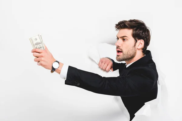 Handsome businessman holding dollar banknotes behind hole in paper wall on white — Stock Photo