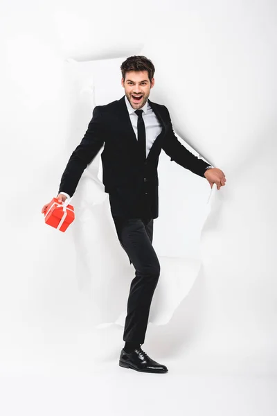 Happy handsome man in suit with present walking through hole in paper on white — Stock Photo