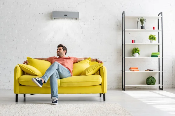 Sonriente hombre guapo sentado en el sofá amarillo bajo el aire acondicionado en casa - foto de stock
