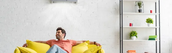 Panoramic shot of handsome man sitting on yellow sofa under air conditioner at home — Stock Photo