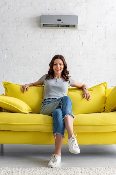 Красива усміхнена жінка сидить на жовтому дивані під кондиціонером вдома — стокове фото