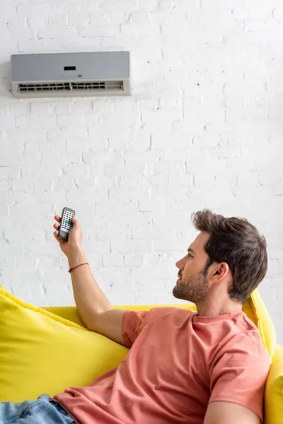 Joven guapo sosteniendo el control remoto mientras está acostado en el sofá bajo el aire acondicionado - foto de stock