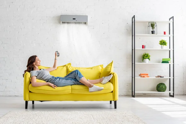 Atractiva joven acostada en el sofá amarillo bajo el aire acondicionado en casa - foto de stock