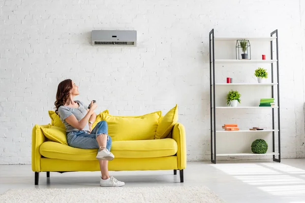 Hübsche junge Frau sitzt zu Hause auf gelbem Sofa unter Klimaanlage — Stockfoto