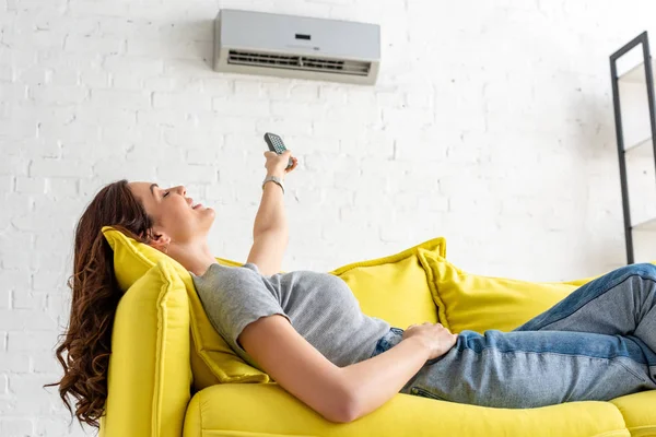 Atraente jovem mulher relaxante sob ar condicionado e segurando controle remoto — Fotografia de Stock