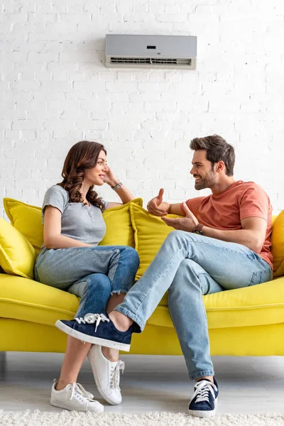 Homme et femme souriants assis sur un canapé jaune sous climatiseur à la maison — Photo de stock