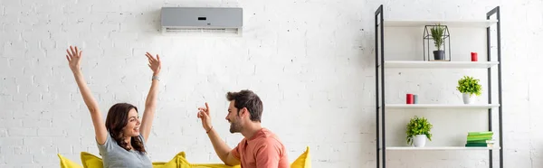 Panoramic shot of excited man and woman sitting under air conditioner at home — Stock Photo