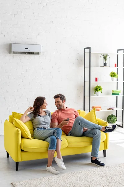 Joven hombre y mujer hablando mientras está sentado en el sofá amarillo bajo el aire acondicionado en casa - foto de stock