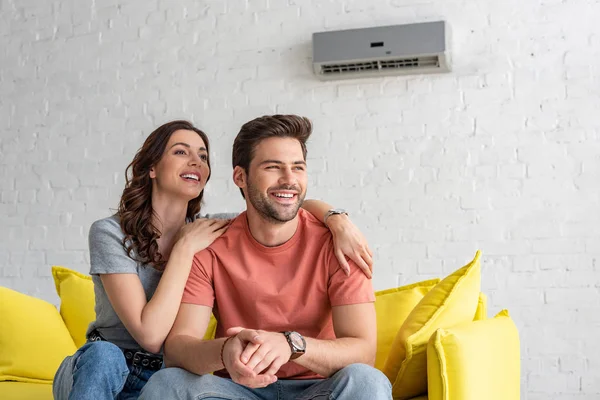 Feliz pareja sentada en el sofá amarillo bajo el aire acondicionado en casa - foto de stock