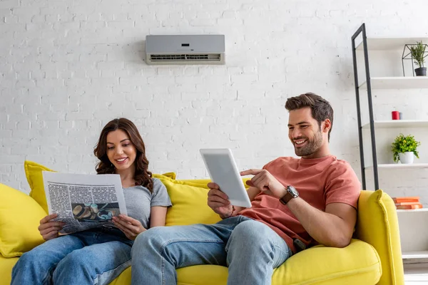 Hübsche Frau beim Zeitungslesen und schöner Mann mit digitalem Tablet, während er zu Hause auf gelbem Sofa unter Klimaanlage sitzt — Stockfoto