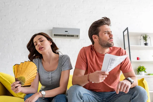 Young woman with hand fan and handsome man with newspaper suffering from heat at home — Stock Photo