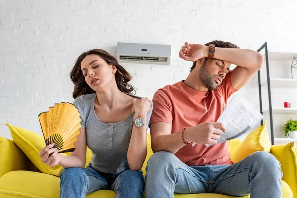 Mujer bonita con ventilador de mano y hombre guapo con periódico que sufre de calor en casa - foto de stock