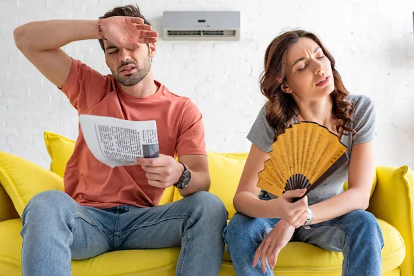 Hombre y mujer exhaustos sentados en el sofá y sufriendo de calor en casa - foto de stock