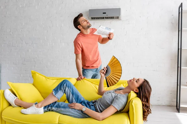 Woman and lying on yellow sofa and man with newspaper suffering from heat at home — Stock Photo