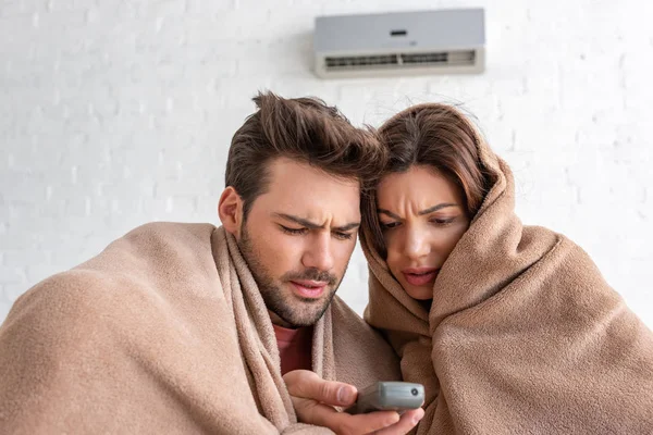 Homme et femme congelés se réchauffant sous des couvertures tout en étant assis sous climatiseur avec télécommande — Photo de stock