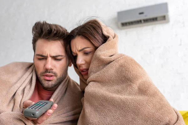 Frozen couple warming under blankets while sitting under air conditioner with remote control — Stock Photo