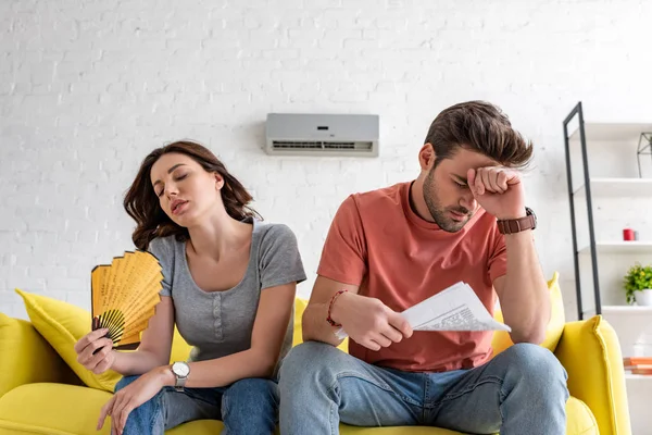Man with newspaper and woman with hand fan sitting on sofa and suffering from heat at home — Stock Photo